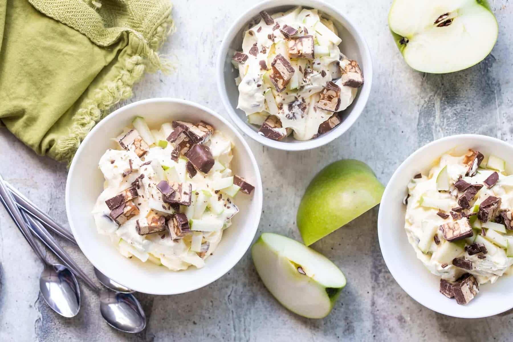 Snickers salad in three bowls surrounded by apple pieces.