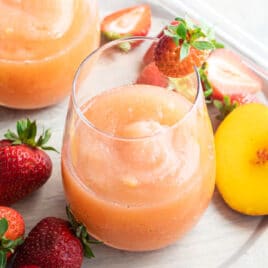 Two glasses of strawberry peach frose on a plate surrounded by slices of strawberries and peaches.