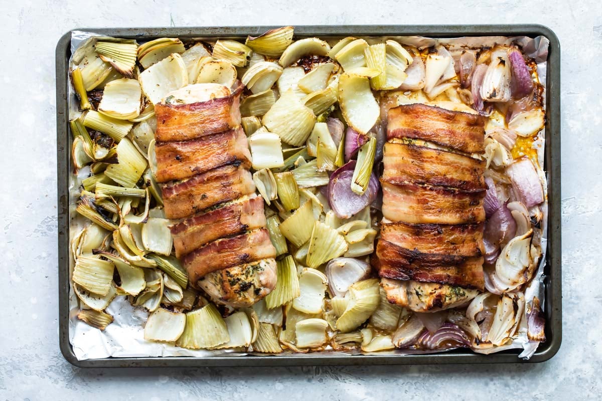 Baked bacon wrapped pork tenderloin on a baking sheet with roasted fennel and onions.
