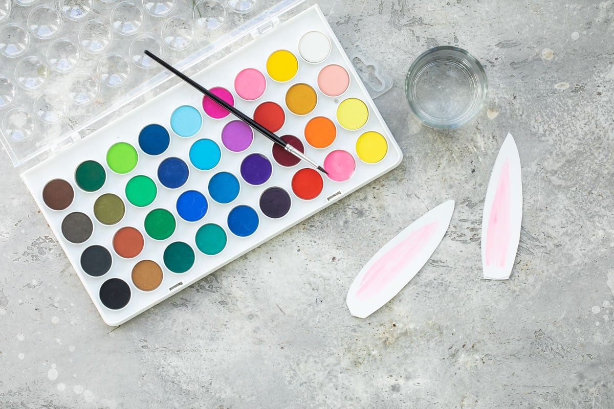 A pallet of water colors, a clear glass with water, and painted bunny ears.