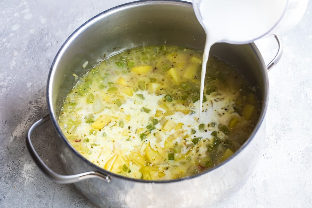 Cream being poured into a silver pot with New England clam chowder ingredients.