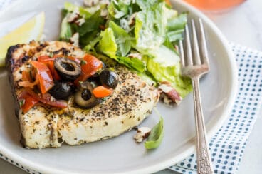 A piece of grilled swordfish on a plate with olive topping and green salad.