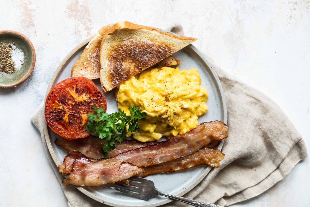 Scrambled eggs on a plate with toast triangles, bacon slices, parsley, and a roasted tomato.