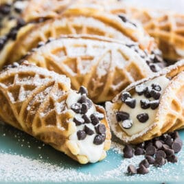Mock italian cannoli with pizzelle on top of a blue cutting board covered in powdered sugar.
