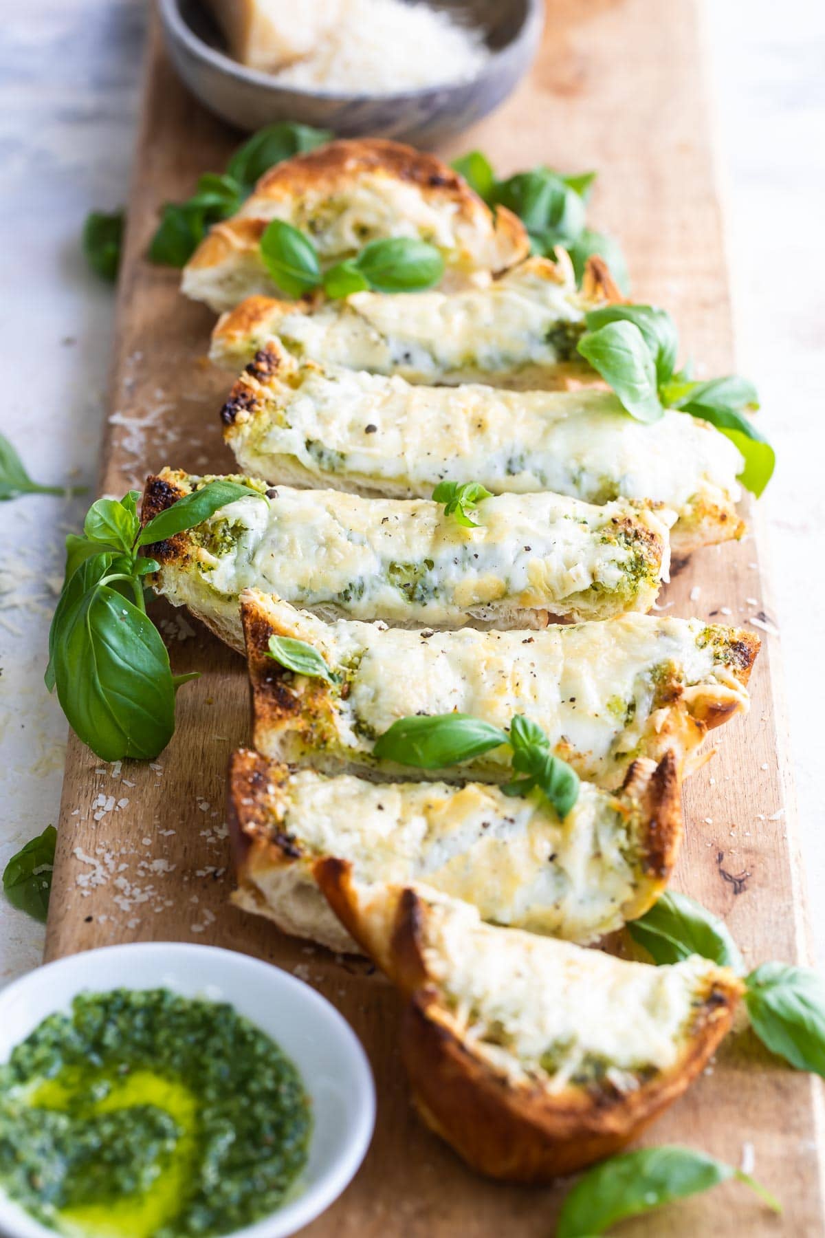 Slices of pesto cheese bread surrounded by basil leaves on a wooden cutting board.