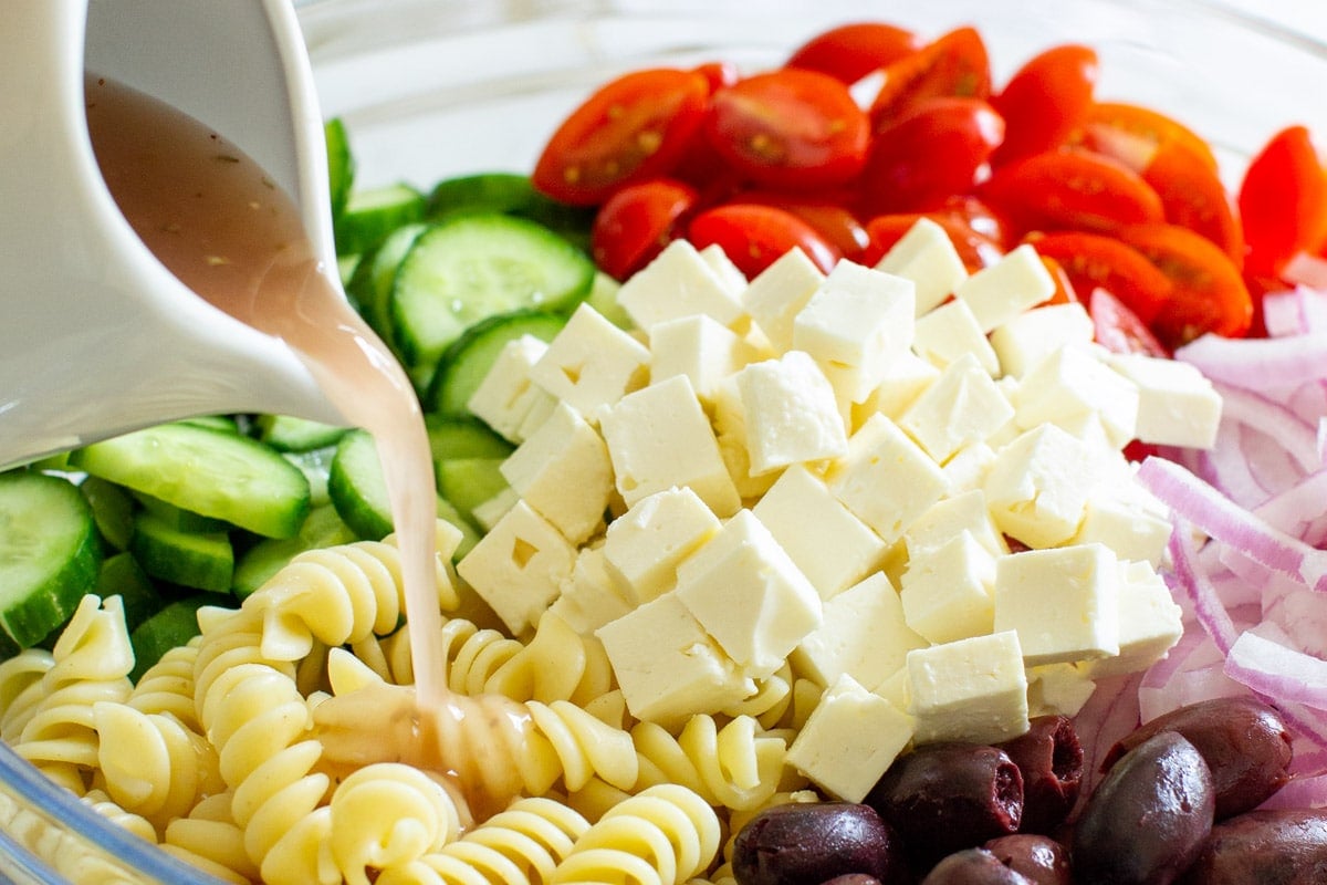 A clear bowl with ingredients prepared to toss for the Greek Pasta Salad, while Homemade Red Wine Vinaigrette is being poured into the bowl from a white serving container.