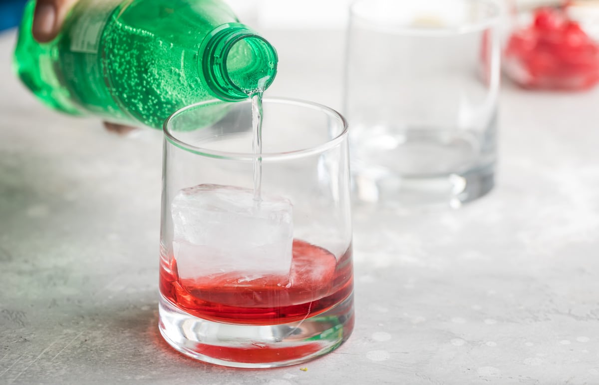 Lemon lime soda being poured over grenadine in a clear glass.