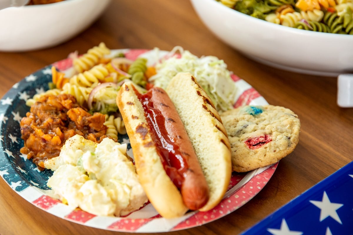 A table of 4th of July Cookout food such as brats, hot dogs, pasta salad, cookies, and drinks.