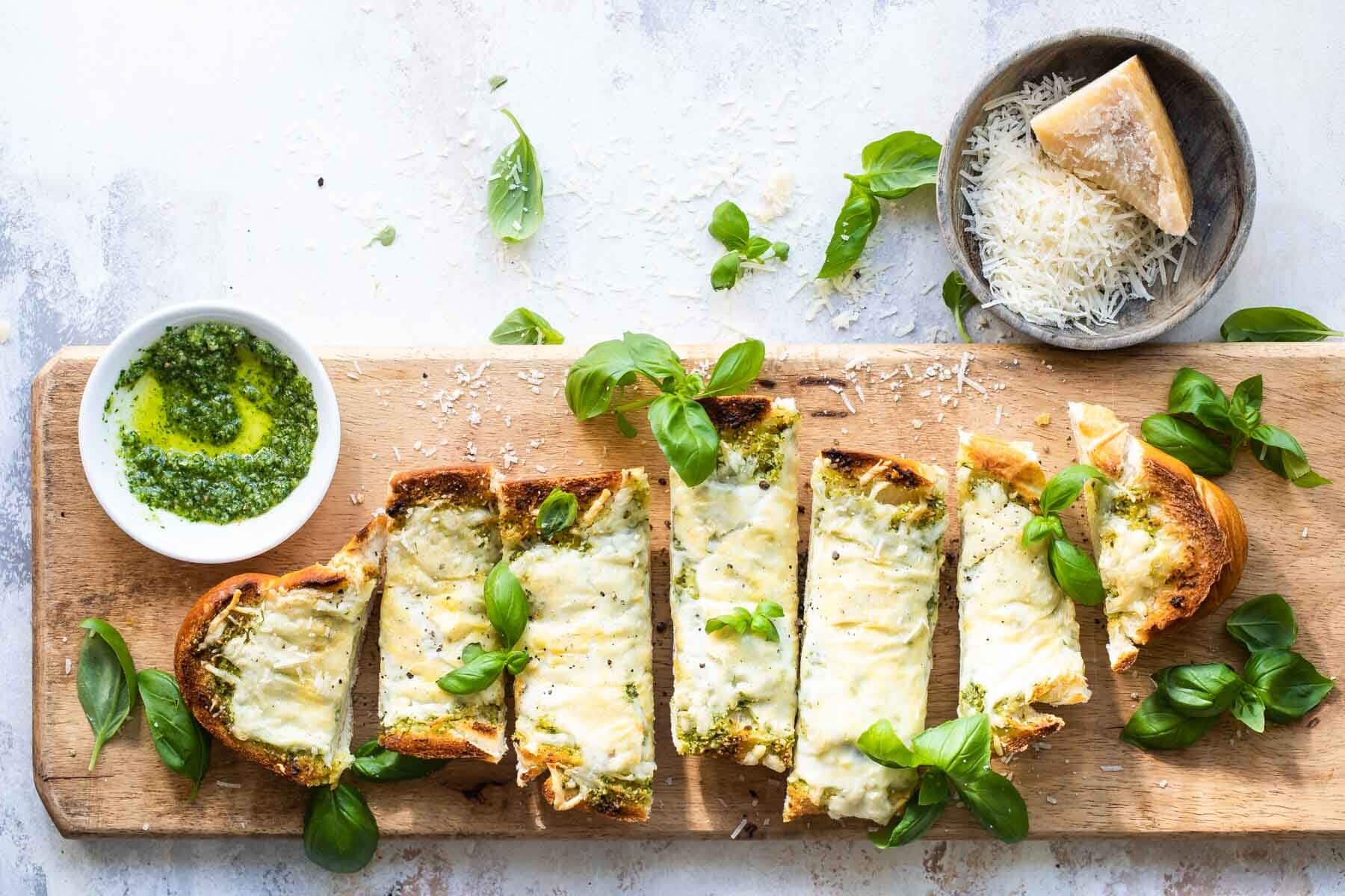 Slices of pesto cheese bread surrounded by basil leaves on a wooden cutting board.