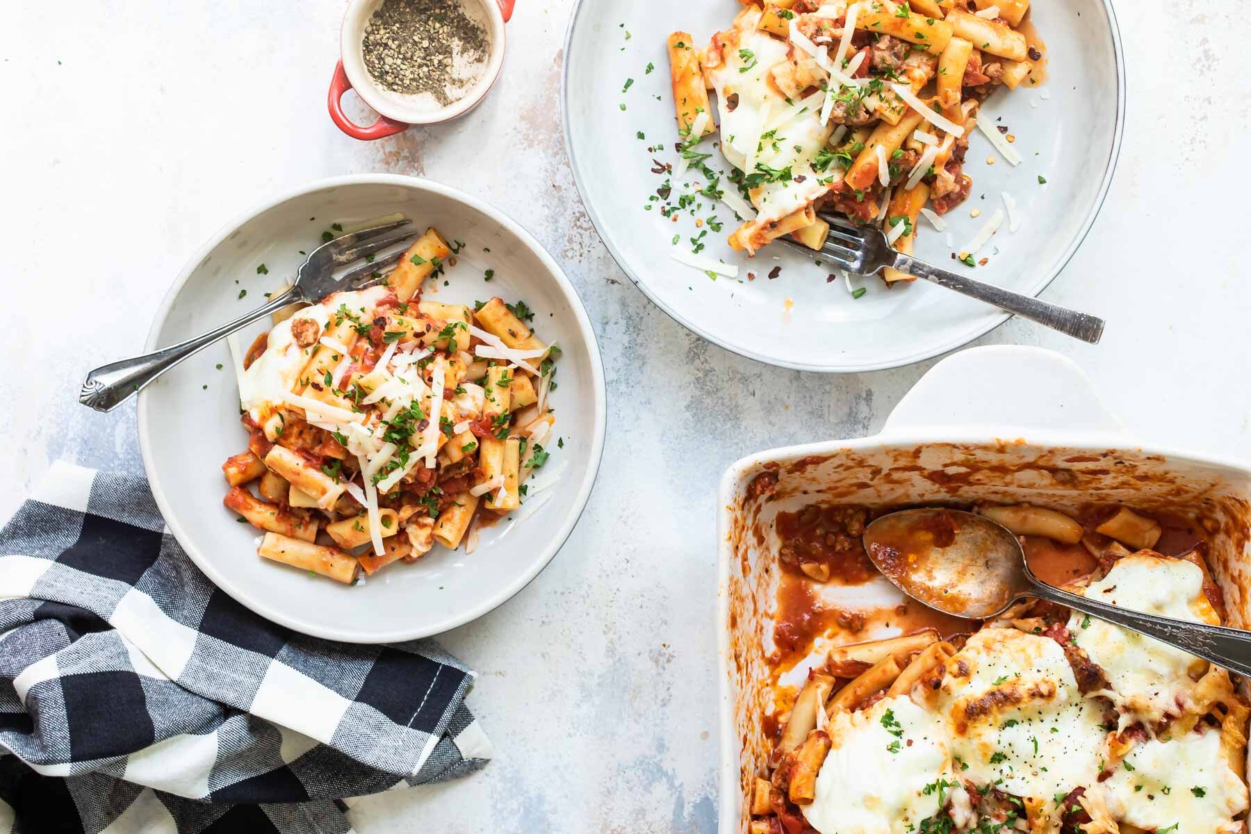 Two plates of baked ziti next to a pan of baked ziti.