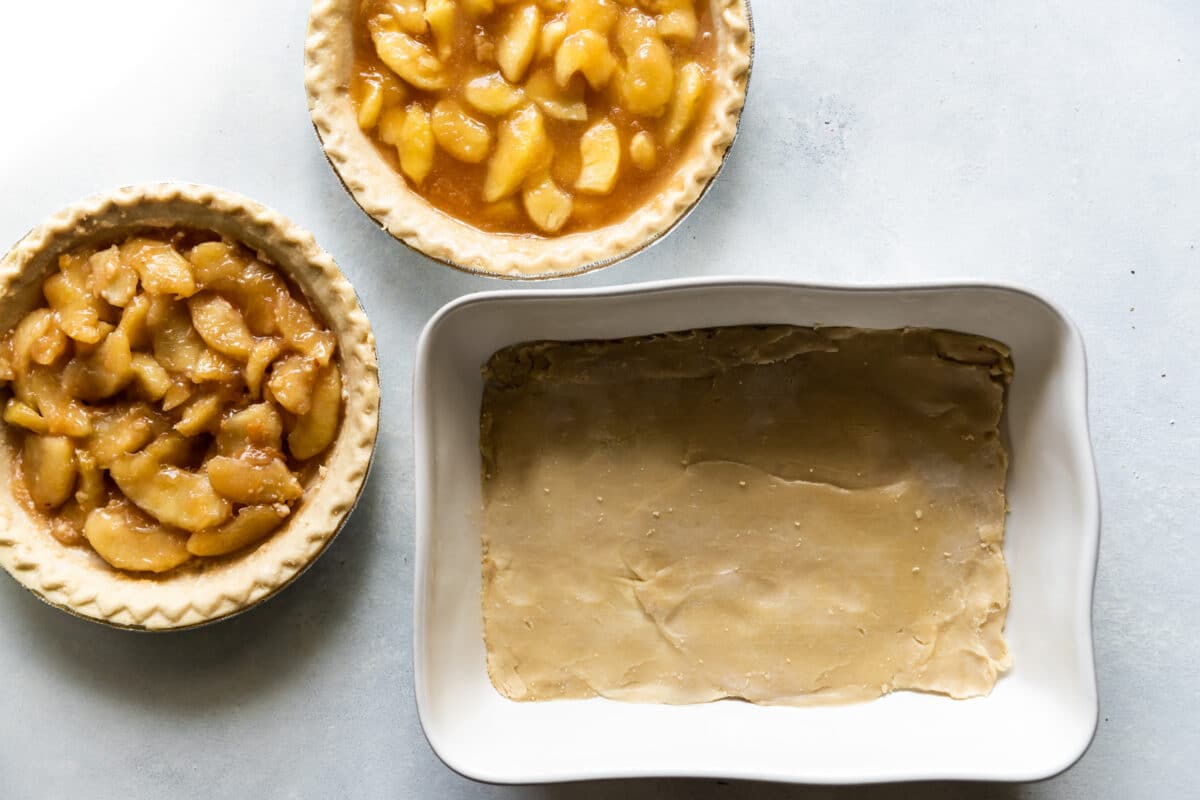 A baking dish with pie crust in the bottom and bowls of apple pie filling next to it.