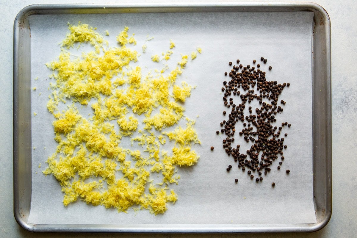 Raw lemon zest and black pepper corns on a baking sheet.
