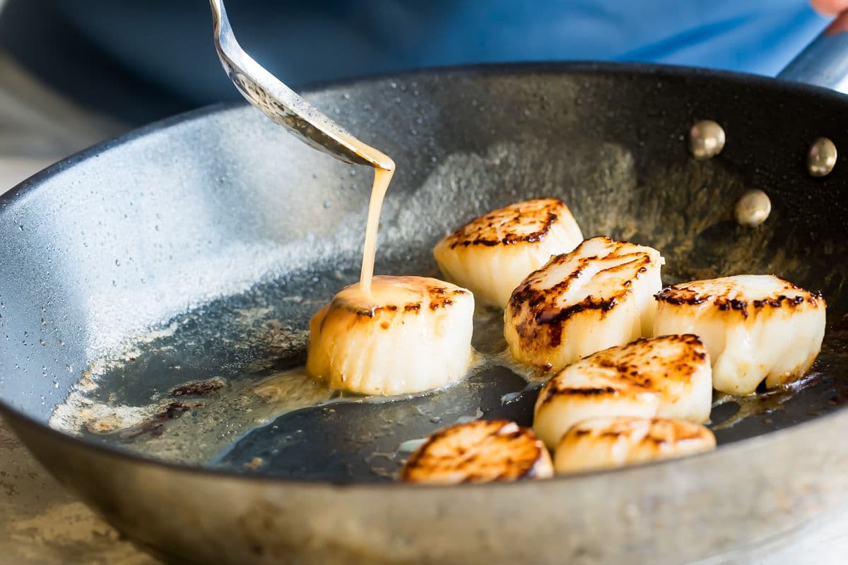 Lemon butter sauce being drizzled onto pan-seared scallops in a black pan.