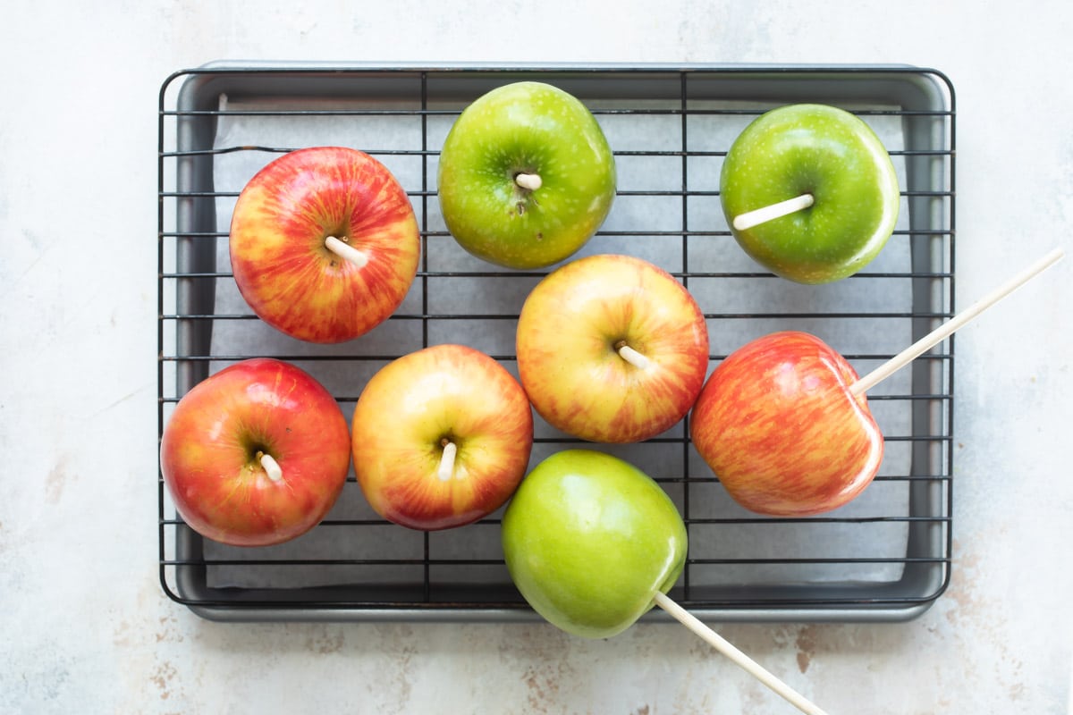 Eight apples with wooden skewers in them resting on a cooling rack.