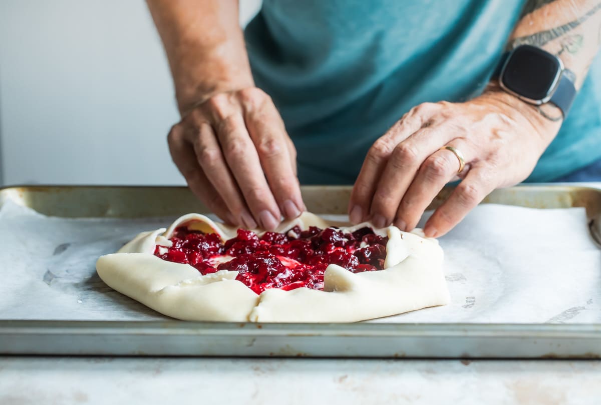 Someone folding an uncooked cranberry cheesecake galette on a piece of parchment paper on a baking sheet.