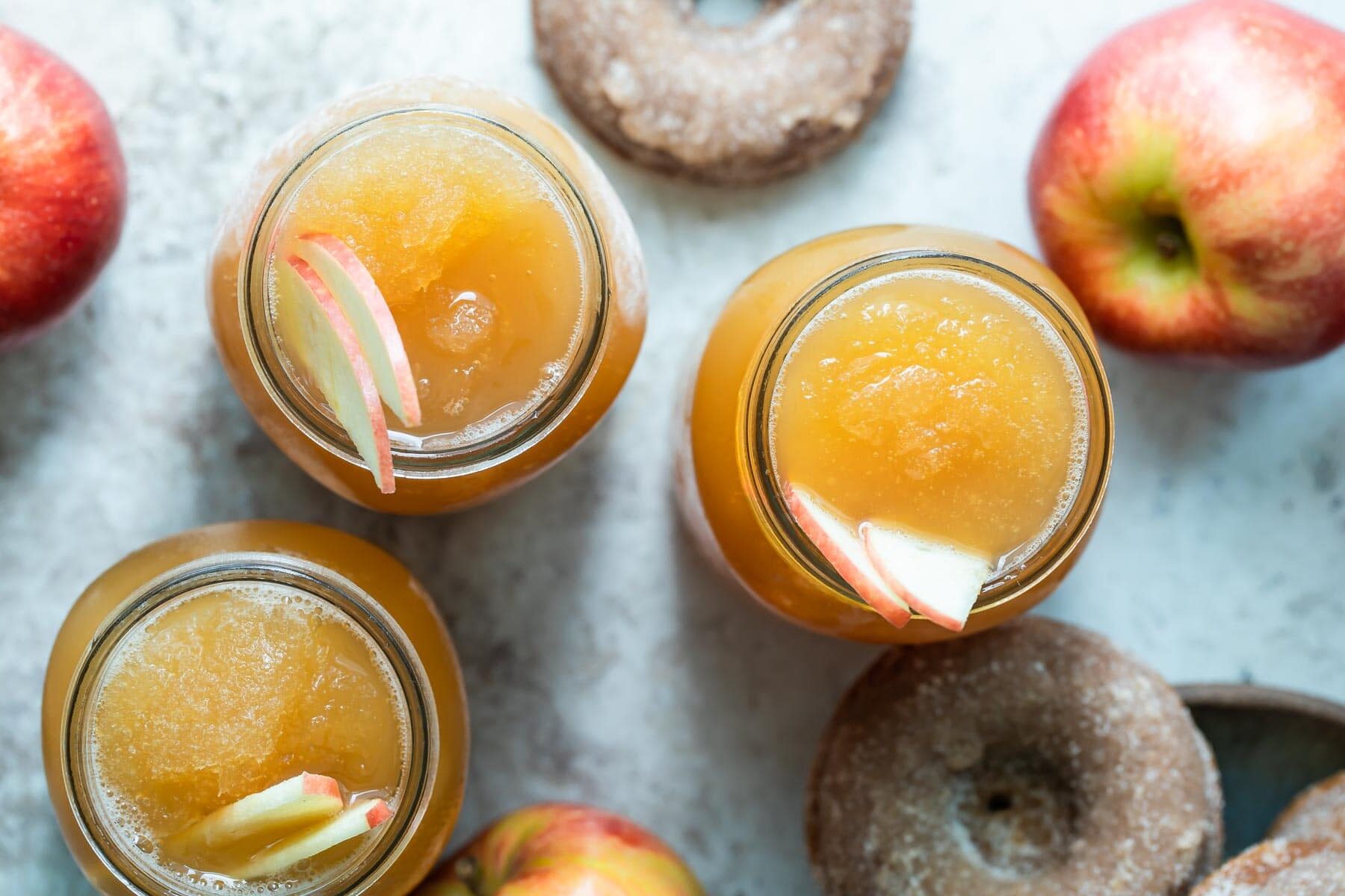 Three mason jars of brandy apple cider slush surrounded by donuts and apples.