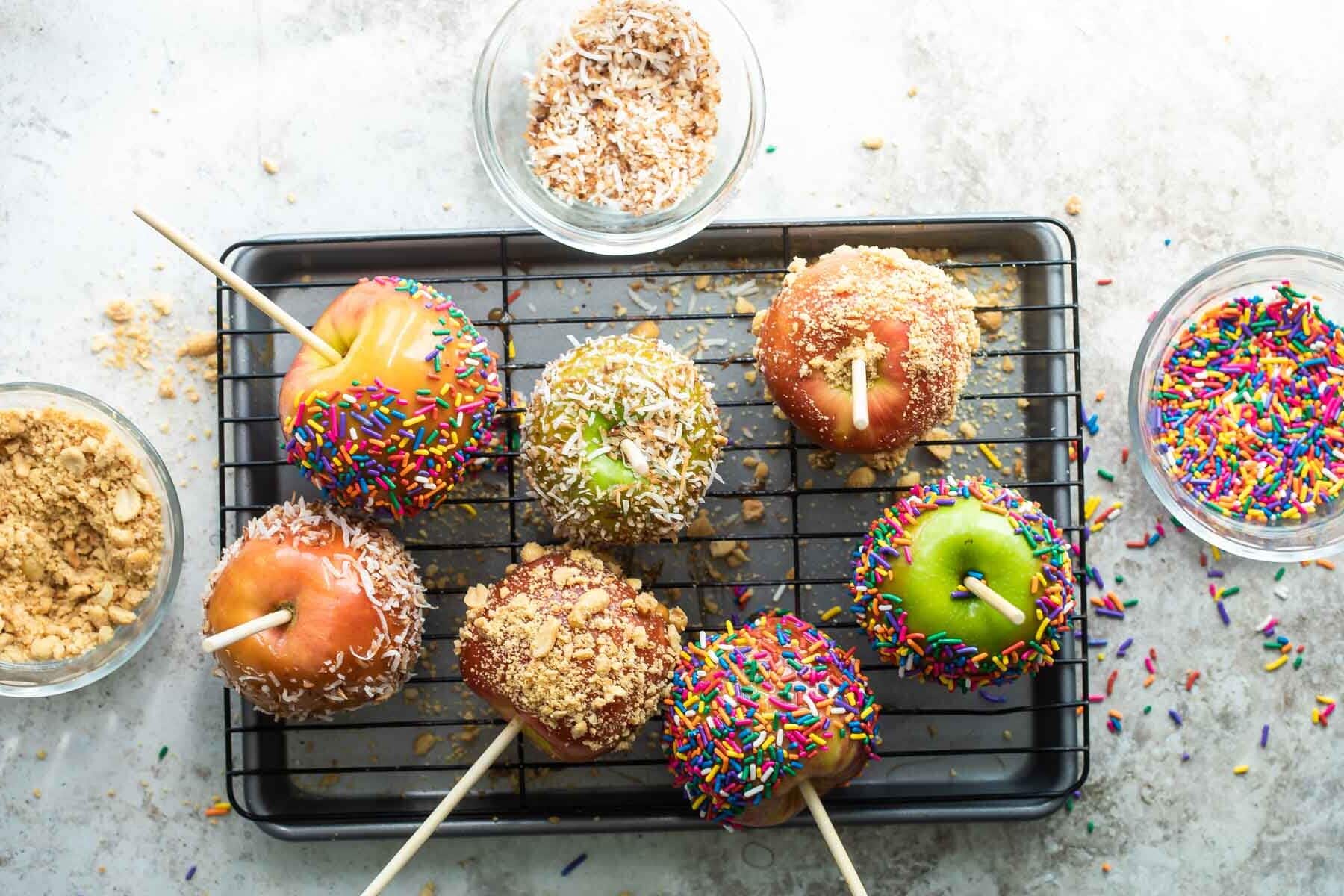 Decorated caramel apples on a cooling rack surrounded by various bowls of toppings.