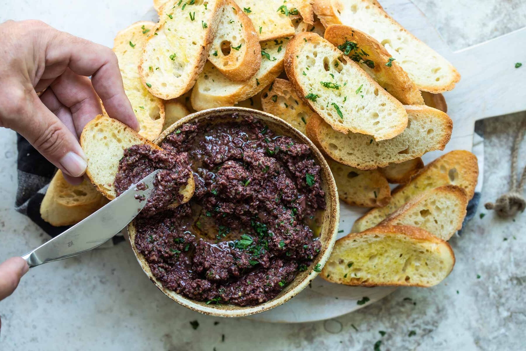 Homemade olive tapenade in a bowl surrounded by toasted bread.