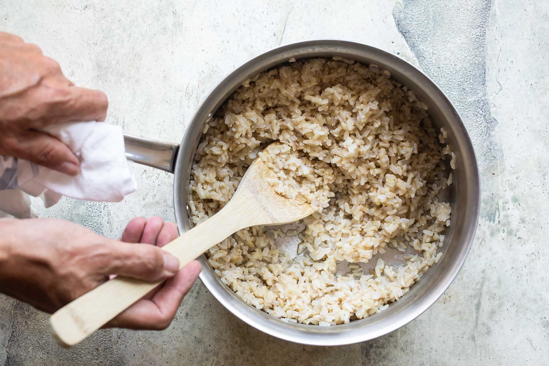 Someone stirring a pot of brown rice with a wooden spoon.