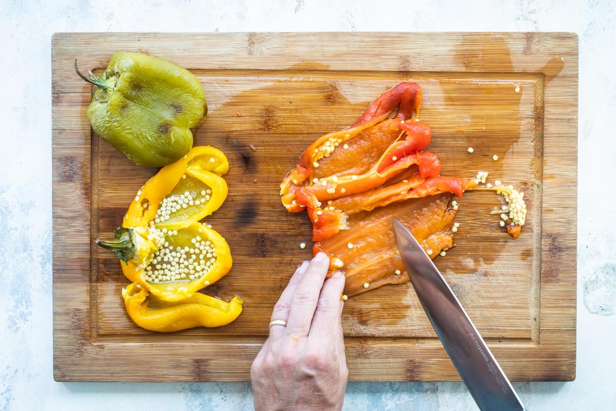 Someone slicing red, yellow, and green roasted peppers on a wood cutting board.