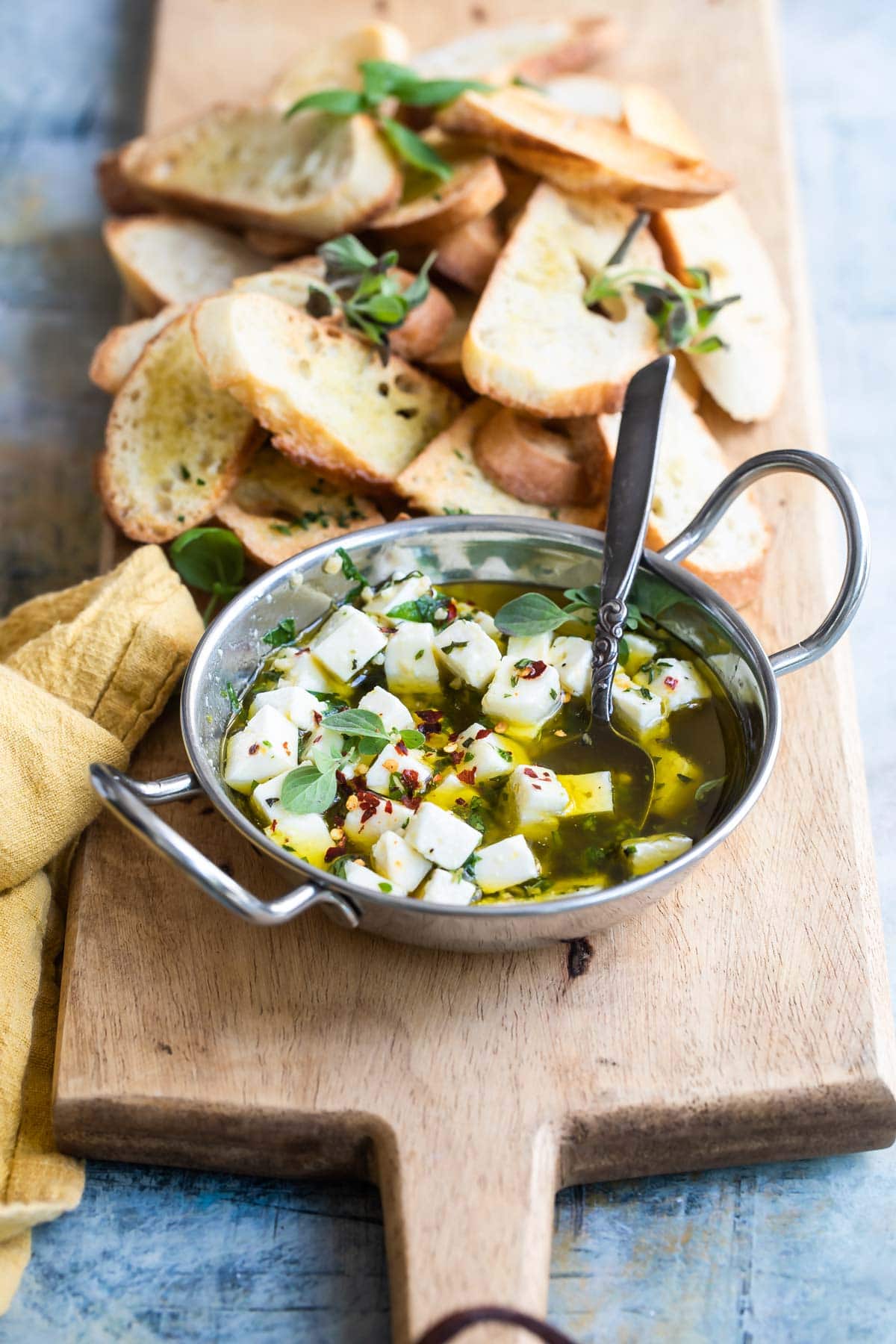 Marinated feta in a silver bowl on a wood board with slices of bread.