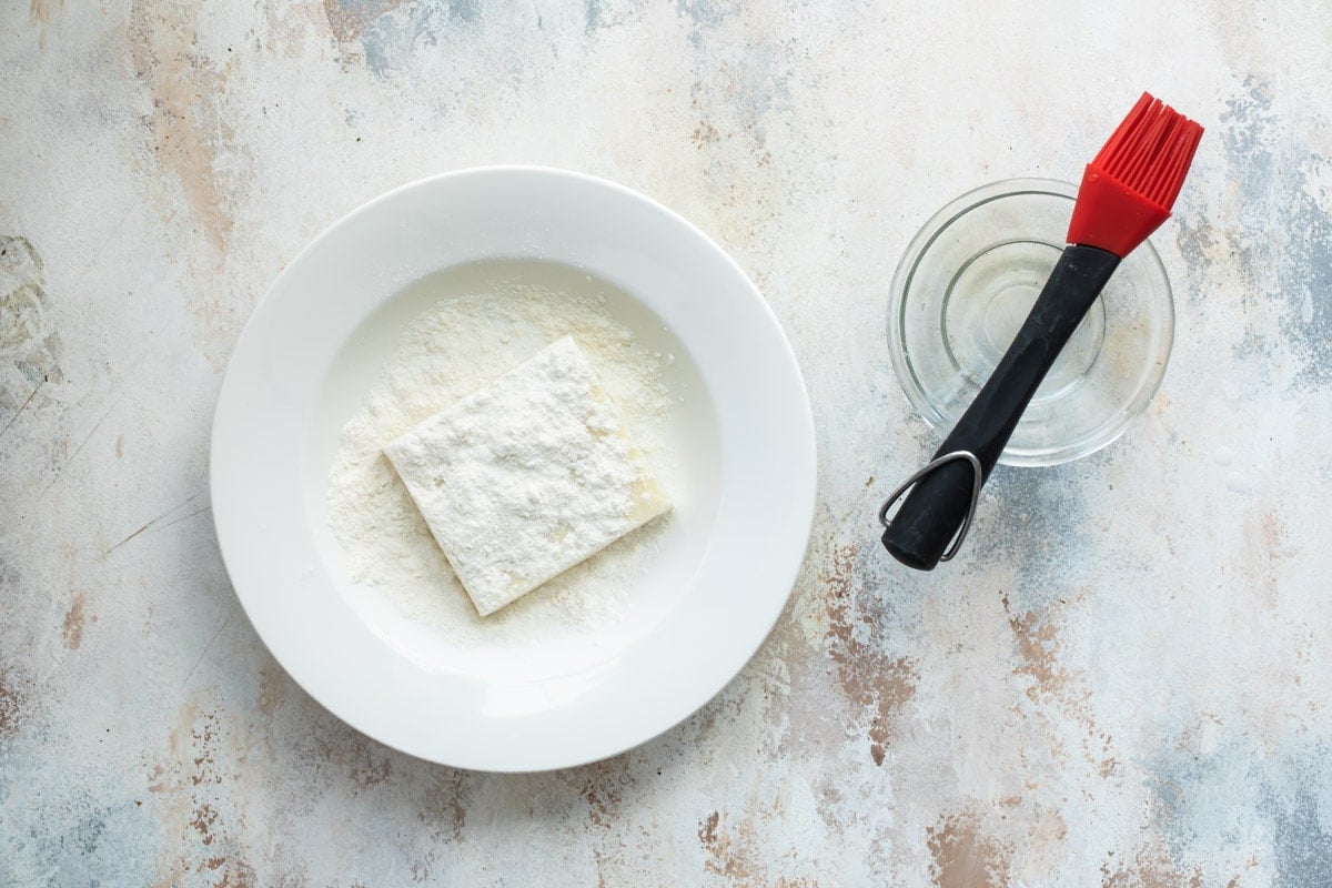 Pastry dough sprinkled with flour on a white plate with a bowl of water and a brush.