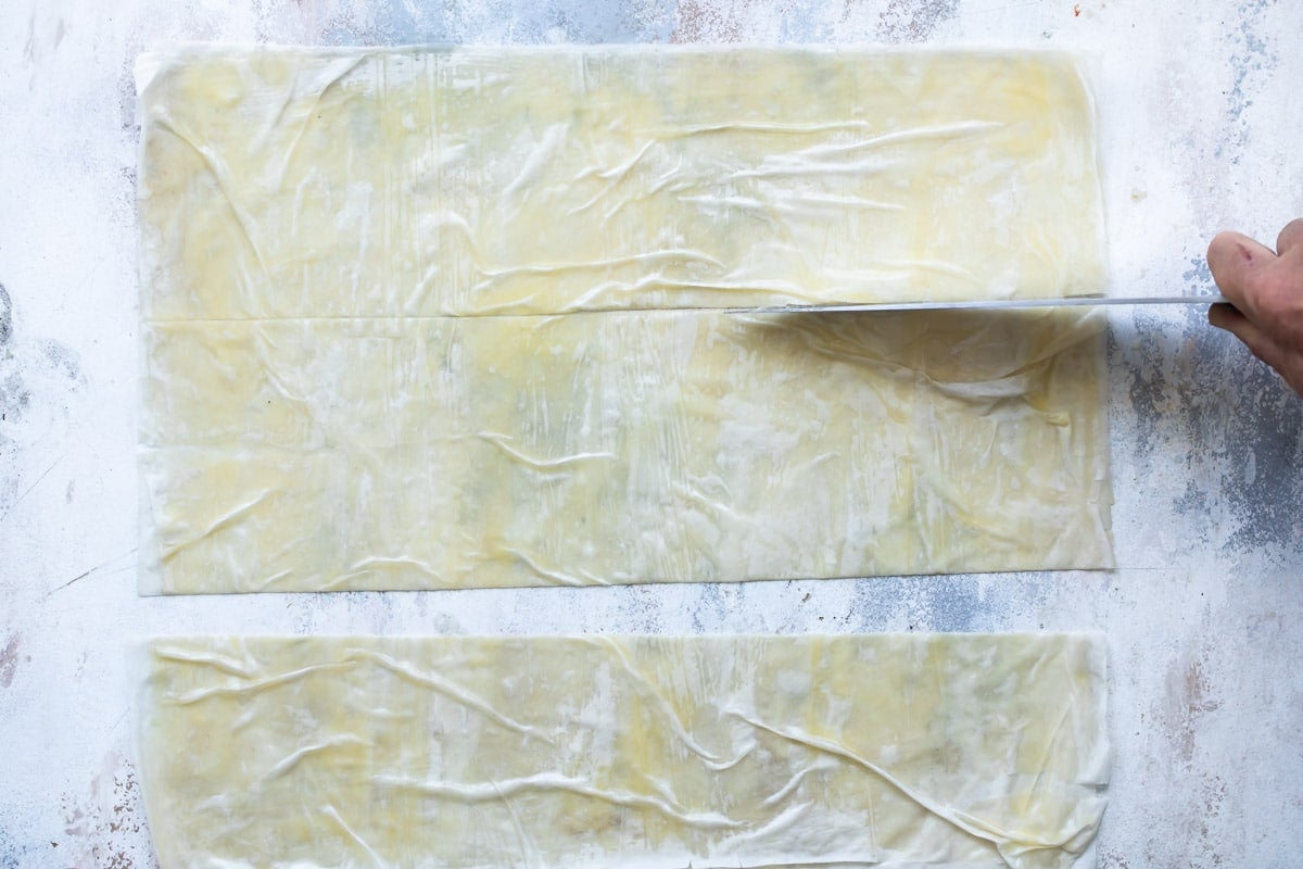 Someone cutting a sheet of pastry dough with a knife into three long rectangles.