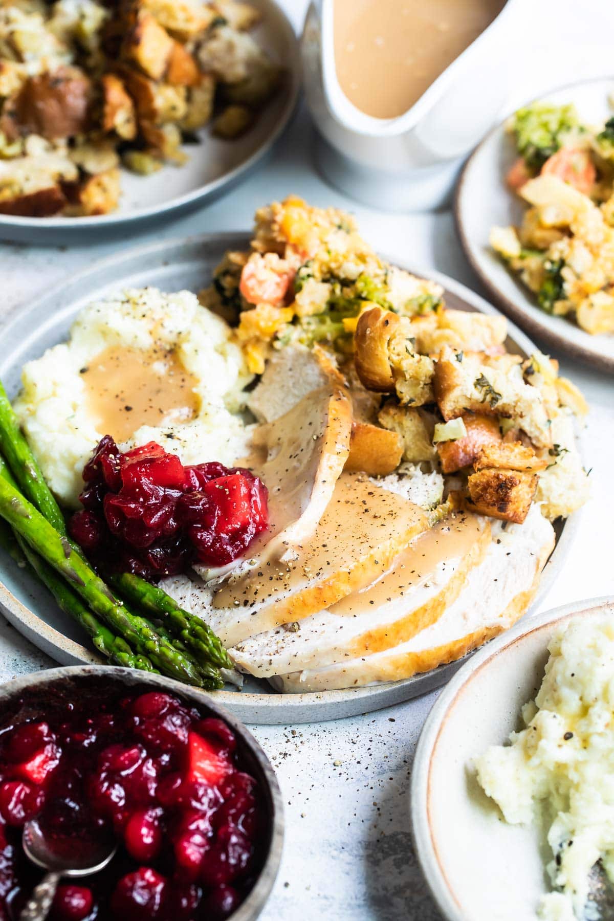 A plate of roasted turkey with gravy, mashed potatoes, bread stuffing, veggie casserole, cranberry sauce with apples, and roasted asparagus.