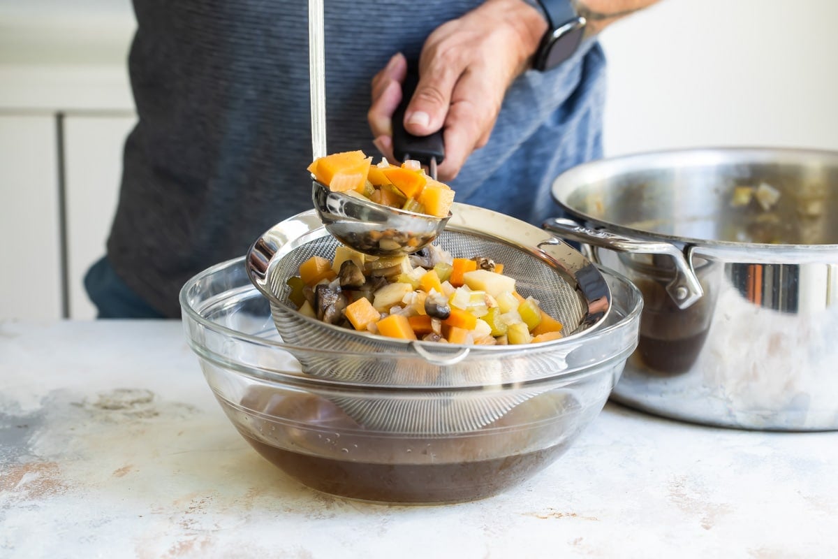 Someone scooping boiled vegetables out of a colander that is resting in a clear bowl filled with vegetable stock.