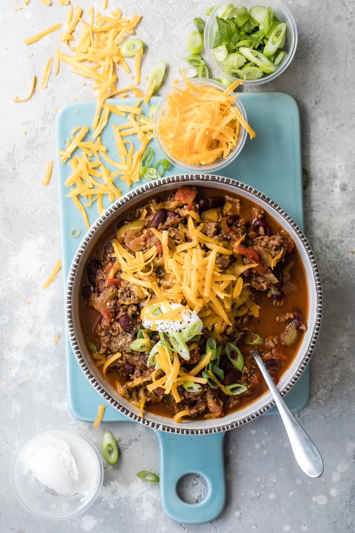 Beef chili in a white bowl on a teal cutting board surrounded by shredded cheese.
