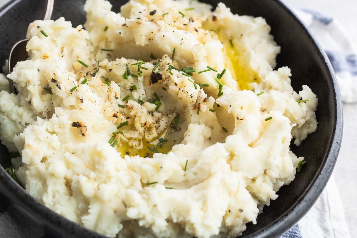 Garlic mashed potatoes in a black bowl with a serving spoon on a blue and white striped towel.