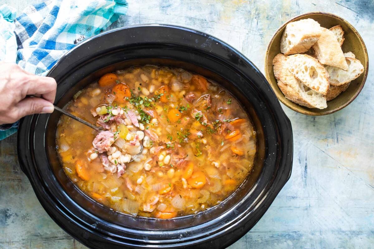 A slow cooker with ham and bean soup being ladled out of it.