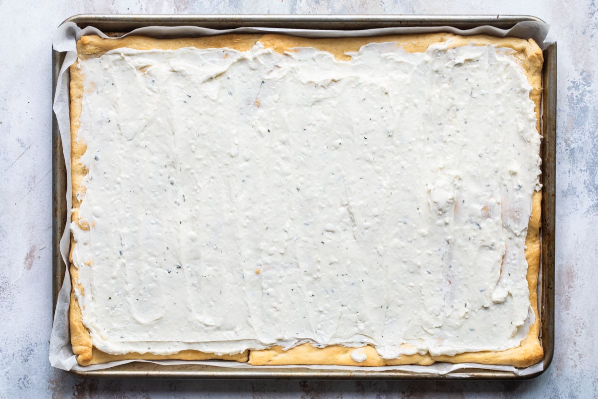 A baked sheet of crescent roll crust topped with veggie pizza dressing on a baking sheet.