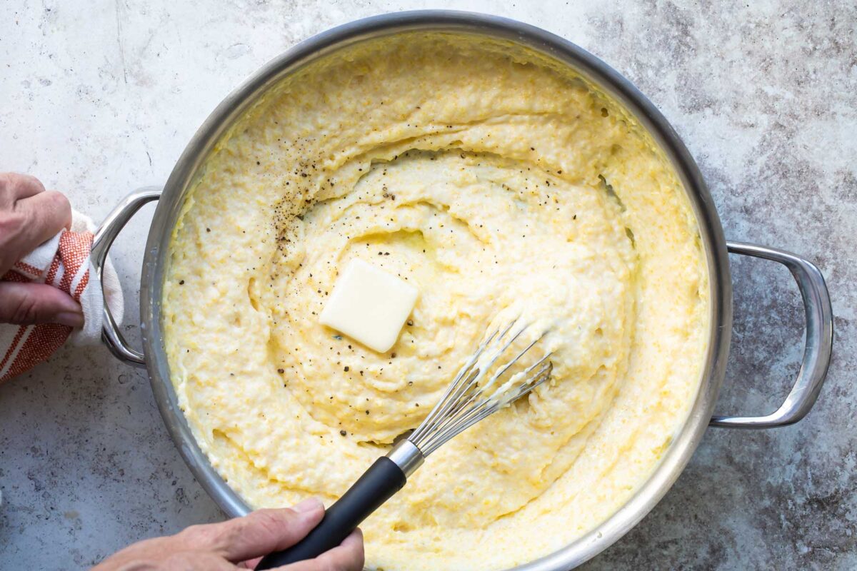 Grits in a silver skillet being stirred with a whisk.