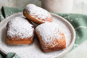 Beignets on a plate next to a coffee mug with liquid in it.