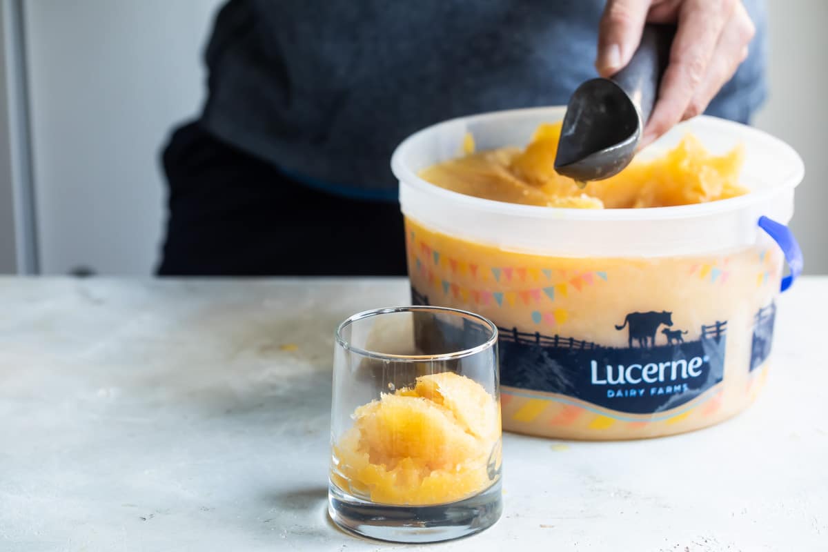 Orange slush being scooped out of an ice cream pail and into a clear glass.
