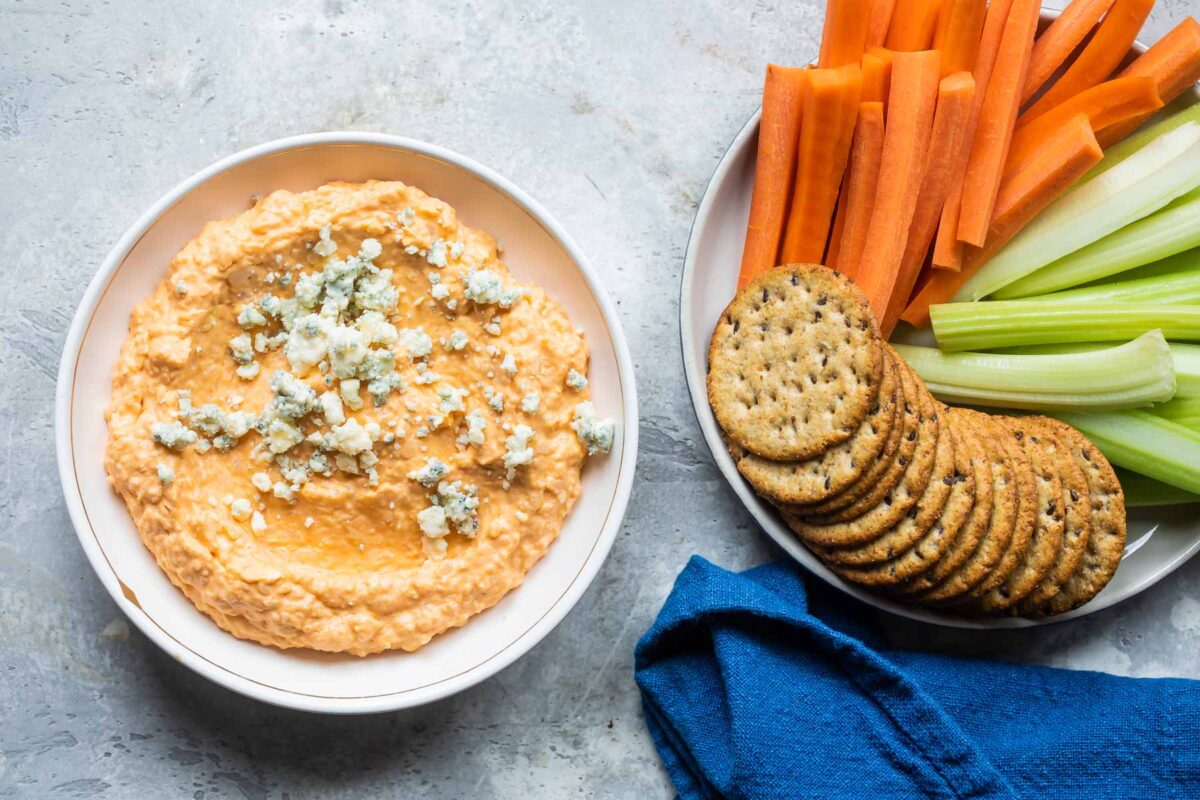 Cauliflower buffalo dip in a bowl next to a bowl of carrots, celery and crackers.