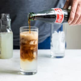 Coke being poured into a half filled glass of lemonade and ice.
