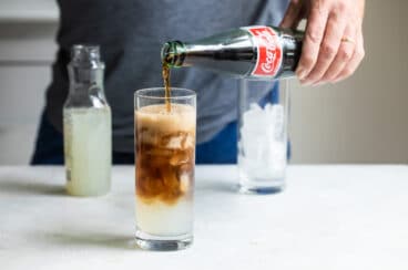 Coke being poured into a half filled glass of lemonade and ice.