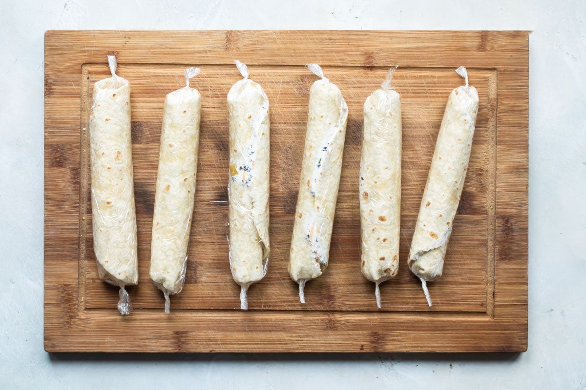 Mexican tortilla roll ups wrapped in plastic wrap on a wooden cutting board.