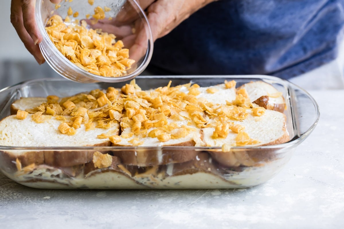 Cereal being poured onto overnight breakfast casserole in a glass baking dish.