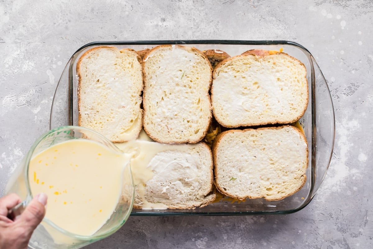 An egg mixture being poured onto overnight breakfast casserole in a glass baking dish.