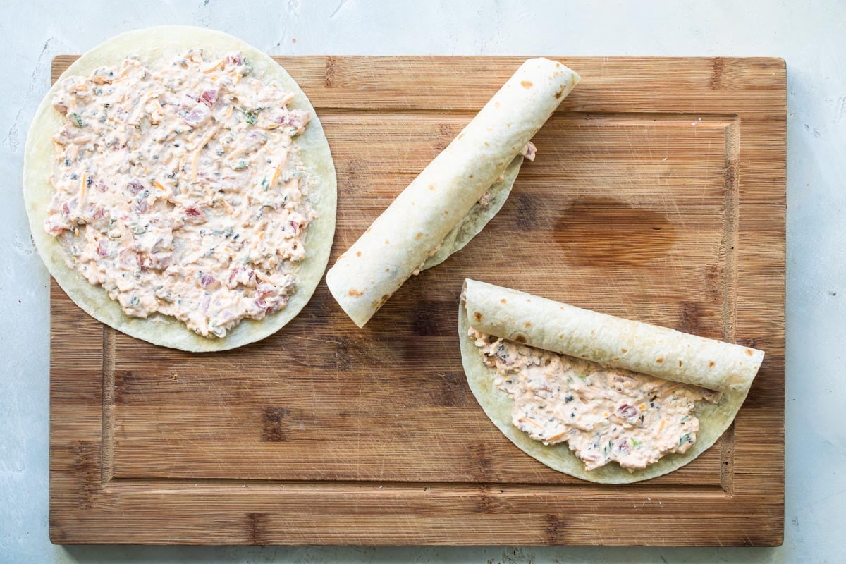Taco tortilla roll ups being rolled on a wooden cutting board