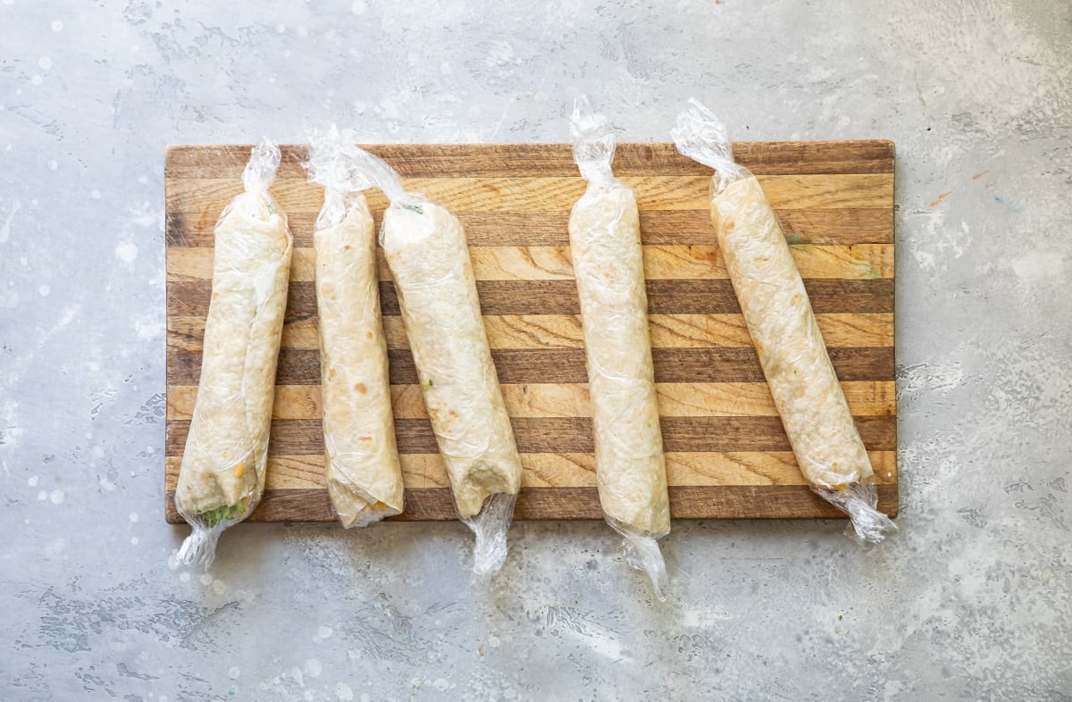 Five vegetable tortilla roll ups wrapped in plastic wrap on a cutting board.