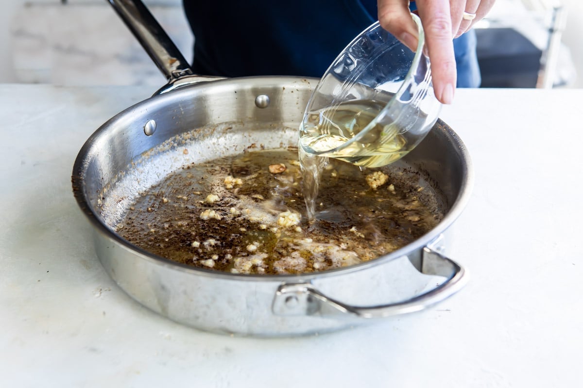 Someone pouring clear liquid into a skillet that was used to cook cajun chicken.