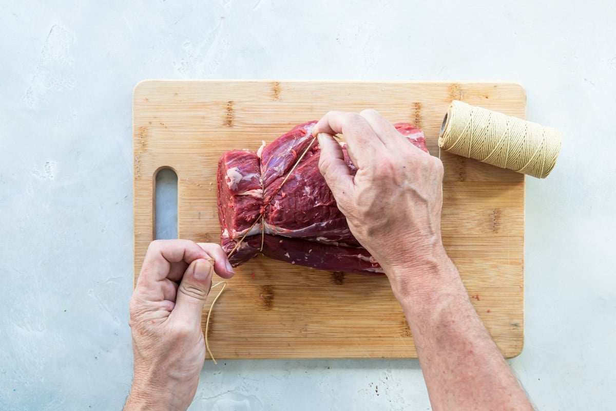 A beef tenderloin being tied on a wooden cutting board.