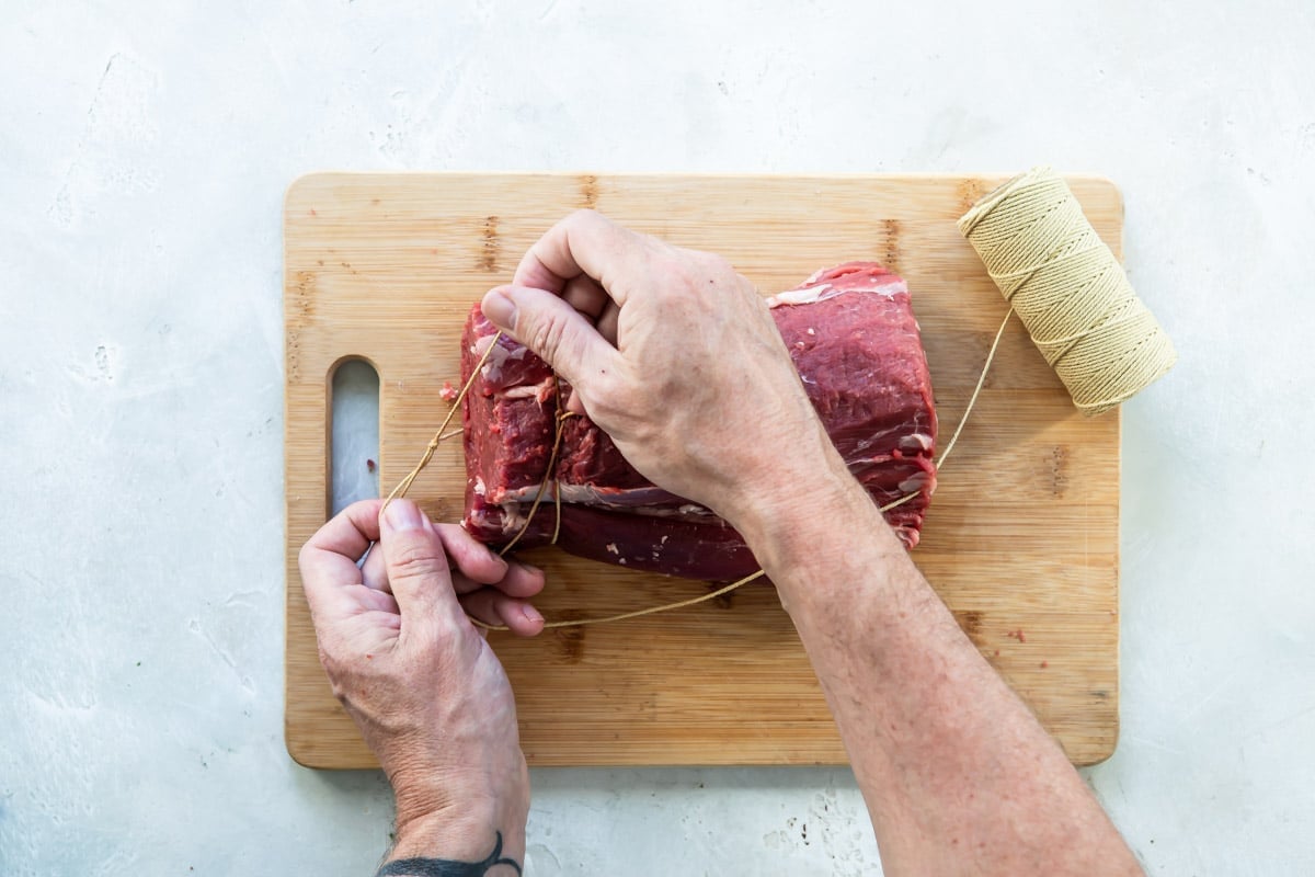 A beef tenderloin being tied on a wooden cutting board.
