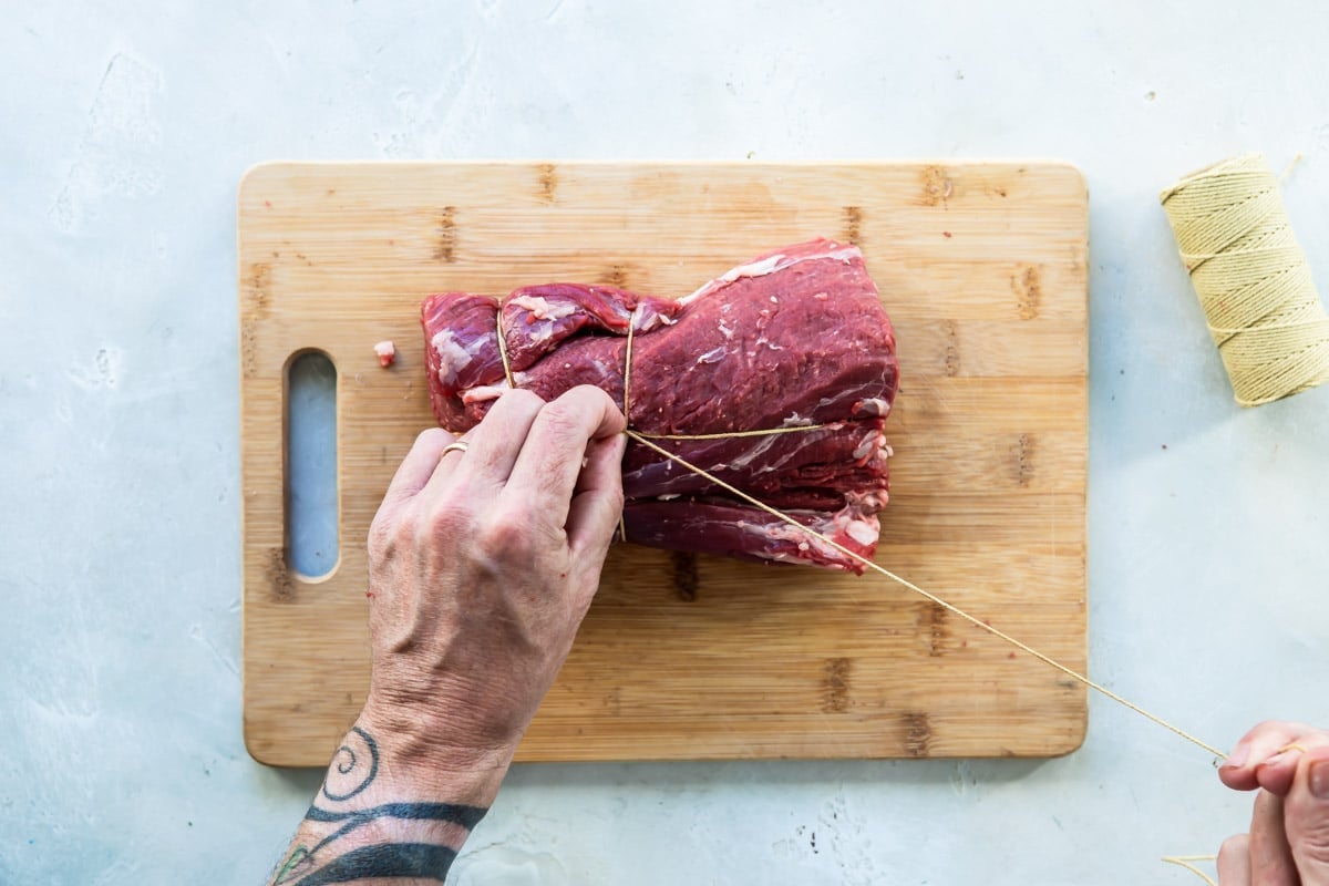 A beef tenderloin being tied on a wooden cutting board.