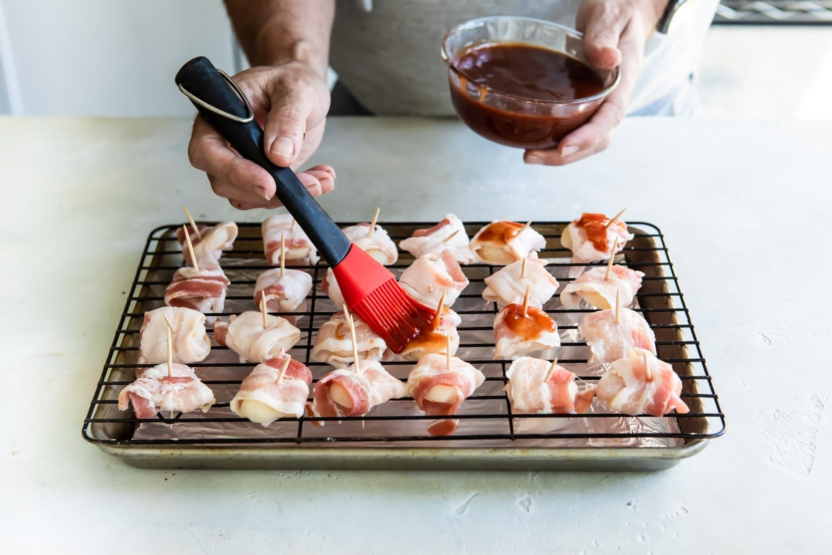 Someone brushing bacon wrapped water chestnuts with barbecue sauce.