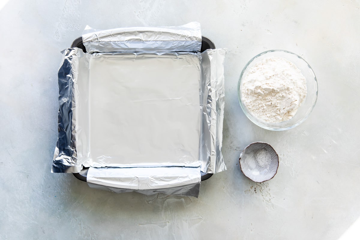 A baking pan with an aluminum foil sling next to two bowls with dry ingredients.